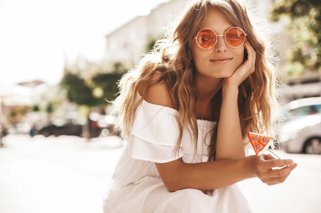 Portrait de beau modèle adolescent blond mignon sans maquillage en été hipster robe blanche vêtements assis sur le fond de la rue