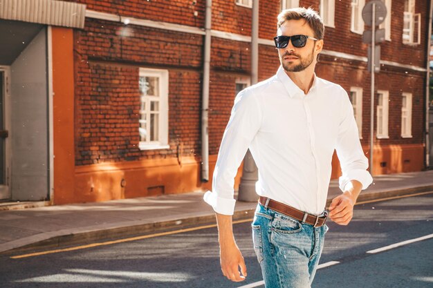 Portrait de beau modèle confiant hipster élégant hipster lambersexuel homme moderne vêtu d'une chemise blanche Mode homme posant sur le fond de la rue en lunettes de soleil