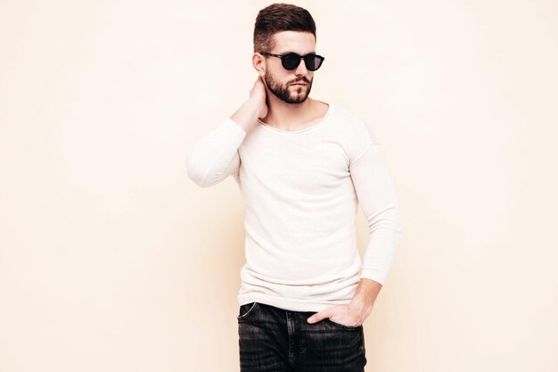 Portrait de beau modèle confiant hipster élégant hipster lambersexuel Homme vêtu d'un pull blanc et d'un jean Mode homme posant près du mur en studio
