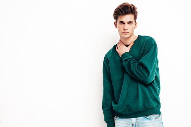 Portrait de beau modèle confiant hipster élégant hipster lambersexuel Homme vêtu d'un pull vert et d'un jean Mode homme posant en studio près d'un mur blanc