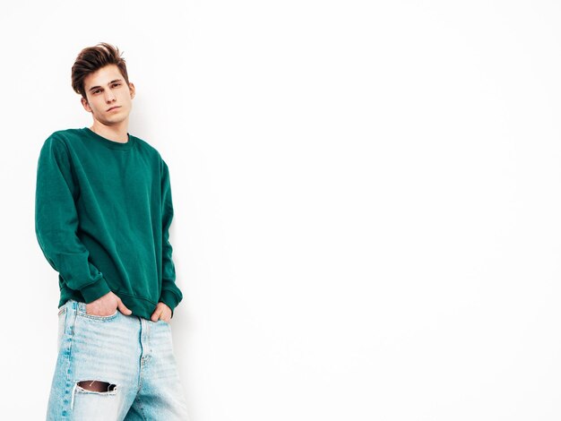 Portrait de beau modèle confiant hipster élégant hipster lambersexuel Homme vêtu d'un pull vert et d'un jean Mode homme posant en studio près d'un mur blanc