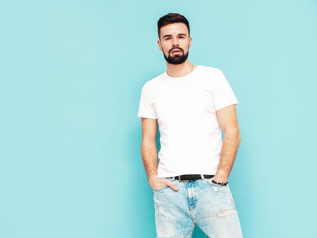 Portrait de beau modèle confiant hipster élégant hipster lambersexuel Homme vêtu d'un t-shirt blanc et d'un jean Mode homme isolé sur mur bleu en studio