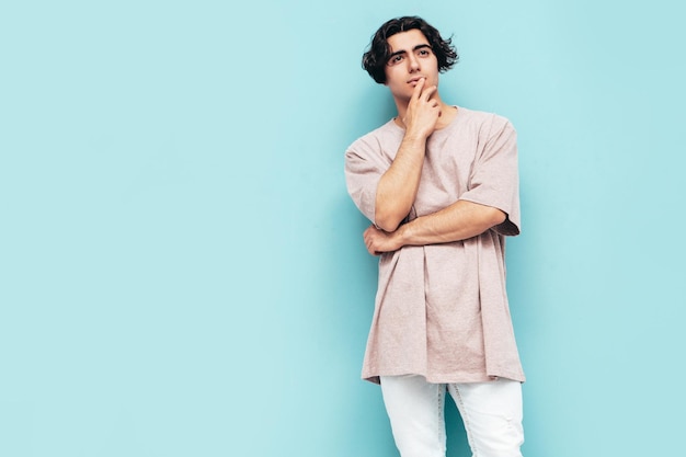 Photo gratuite portrait de beau modèle confiant hipster élégant hipster lambersexuel homme vêtu d'un t-shirt et d'un jean surdimensionnés homme de mode isolé en studio posant près du mur bleu