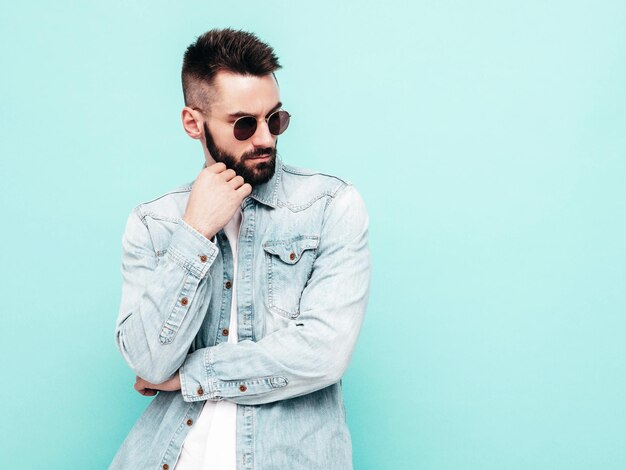 Portrait de beau modèle confiant hipster élégant hipster lambersexuel Homme vêtu d'une veste et d'un jean Mode homme posant près du mur bleu en studio en lunettes de soleil Réfléchi