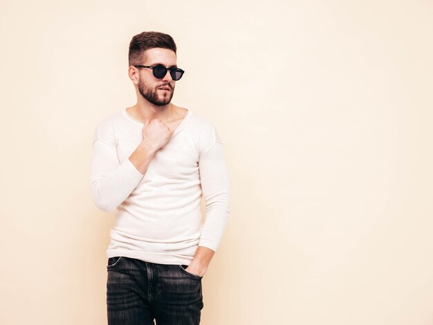 Portrait de beau modèle confiant hipster élégant hipster lambersexuel vêtu d'un pull blanc et d'un jean Mode homme posant près du mur en studio