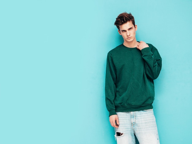 Portrait de beau modèle confiant hipster élégant hipster lambersexuel vêtu d'un pull vert et d'un jean Mode homme posant en studio près du mur bleu
