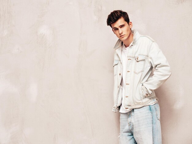 Portrait de beau modèle confiant hipster élégant hipster lambersexuel vêtu d'une veste et d'un jean Mode homme posant en studio près d'un mur gris