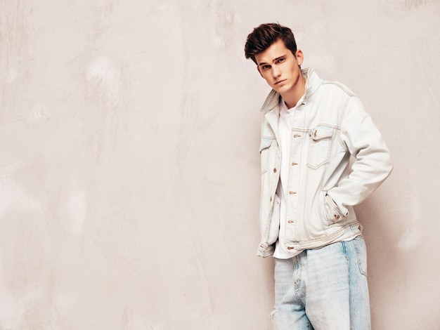 Portrait de beau modèle confiant hipster élégant hipster lambersexuel vêtu d'une veste et d'un jean Mode homme posant en studio près d'un mur gris