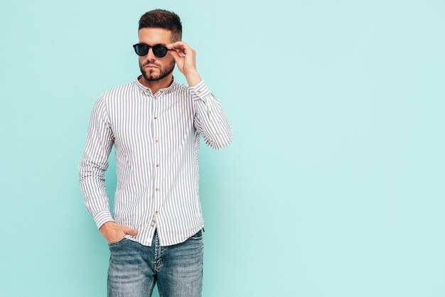 Portrait de beau modèle confiant Sexy homme élégant vêtu d'une chemise et d'un jean Mode hipster mâle posant près du mur bleu en studio Enthousiaste et heureux En lunettes de soleil