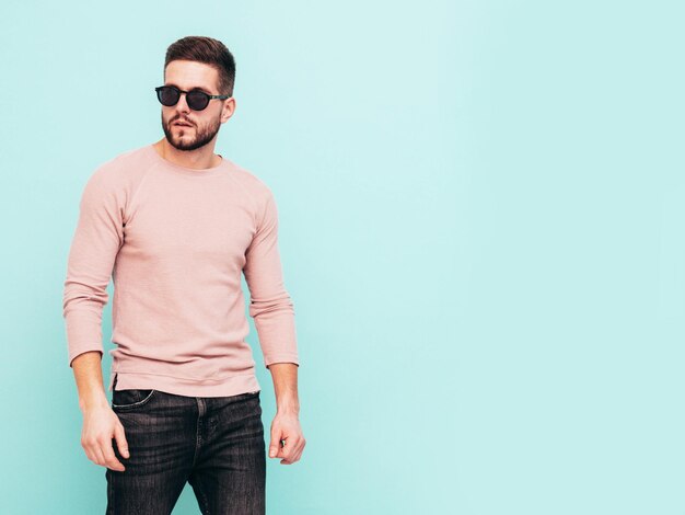 Portrait de beau modèle confiant Sexy homme élégant vêtu d'un pull rose et d'un jean Mode hipster mâle posant près du mur bleu en studio En lunettes de soleil Isolé