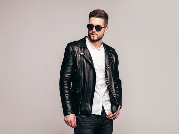 Portrait de beau modèle confiant Sexy homme élégant vêtu d'une veste en cuir de motard et d'un jean noir Mode hipster mâle isolé sur fond gris en studio à lunettes de soleil Isolé