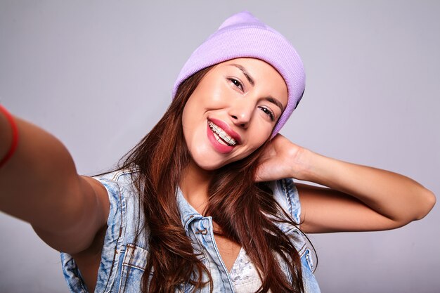 Portrait de beau modèle de femme brune mignonne souriante dans des vêtements de jeans d'été décontractés sans maquillage en bonnet violet faisant photo selfie sur téléphone isolé sur gris