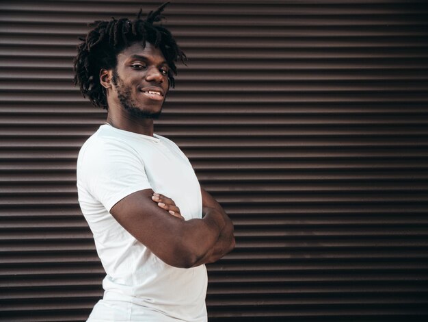 Portrait d'un beau modèle hipster souriant Un homme africain mal rasé vêtu d'un t-shirt d'été blanc et d'un jean Mode masculine avec une coiffure de dreadlocks posant sur le fond de la rue