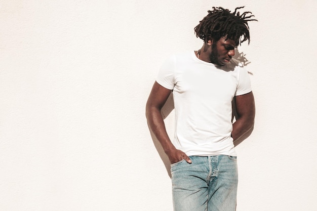 Portrait d'un beau modèle hipster souriant Un homme africain mal rasé vêtu d'un t-shirt d'été blanc et d'un jean Mode masculine avec une coiffure de dreadlocks posant sur le fond de la rue