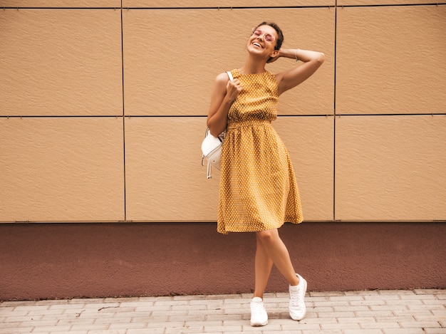 Portrait De Beau Modèle Hipster Souriant Vêtu D'une Robe D'été Jaune. Fille Branchée Posant Dans La Rue. Femme Drôle Et Positive S'amuser