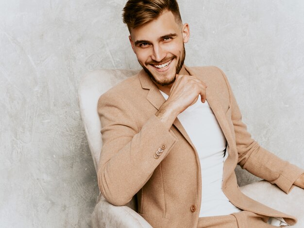 Portrait de beau modèle d'homme d'affaires souriant hipster portant un costume beige décontracté. assis sur une chaise à l'intérieur