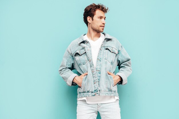 Portrait de beau modèle lambersexuel hipster élégant et confiant avec une coiffure frisée Homme sexy vêtu d'une veste en jean Mode homme isolé sur mur bleu en studio