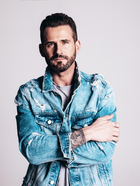 Portrait de beau modèle lambersexuel hipster élégant et confiant Homme sexy vêtu d'une veste en jean Homme de mode isolé sur fond gris en studio