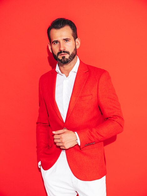 Portrait de beau modèle lambersexuel hipster élégant et confiant Sexy homme moderne vêtu d'un costume élégant Mode homme posant en studio près du mur rouge