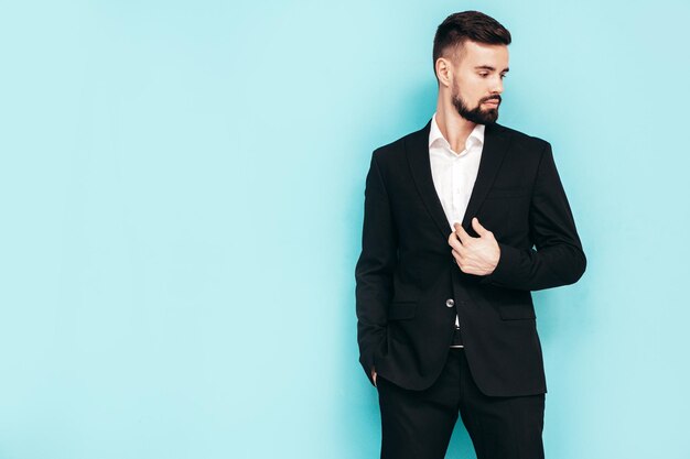 Portrait de beau modèle lambersexuel hipster élégant et confiant Sexy homme moderne vêtu d'un élégant costume noir Mode homme posant en studio près du mur bleu