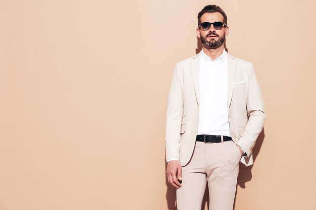 Portrait de beau modèle lambersexuel hipster élégant et souriant Sexy homme moderne vêtu d'un costume blanc élégant Homme de mode avec une coiffure bouclée qui pose en studio près d'un mur beige