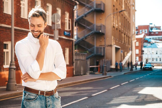 Portrait de beau modèle sexuel hipster élégant et souriant Homme moderne vêtu d'une chemise blanche Mode homme pensif posant sur le fond de la rue à l'extérieur au coucher du soleil