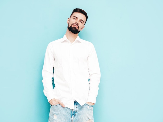 Portrait de beau modèle souriant Sexy homme élégant vêtu d'une chemise blanche et d'un jean Mode hipster mâle posant près du mur bleu en studio Isolé