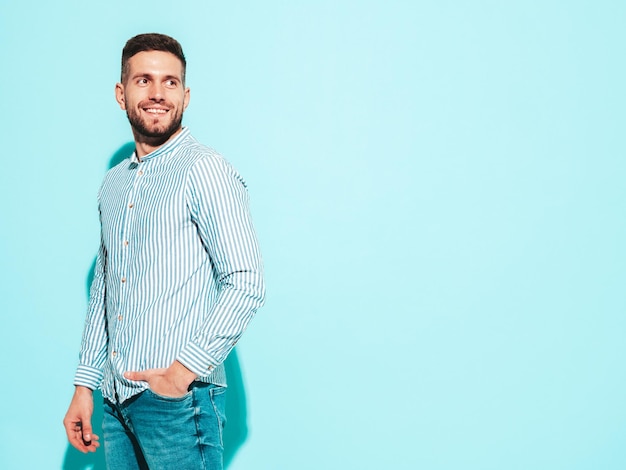 Portrait de beau modèle souriant Sexy homme élégant vêtu d'une chemise et d'un jean Mode hipster mâle posant près du mur bleu en studio Enthousiaste et heureux