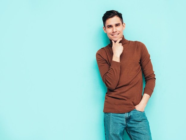 Portrait de beau modèle souriant Sexy homme élégant vêtu d'un pull à col roulé et d'un jean Mode hipster mâle posant sur fond gris en studio Isolé Touchant son menton