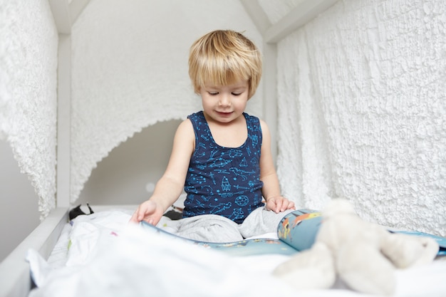 Portrait de beau petit garçon de race blanche aux cheveux blonds habillé en pyjama assis sur un lit à baldaquin blanc, absorbé dans la lecture de livre pour enfants, regardant à travers des images avec une expression intéressée