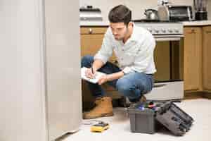 Photo gratuite portrait d'un beau technicien masculin faisant un rapport de travail sur un réfrigérateur cassé dans une cuisine à domicile