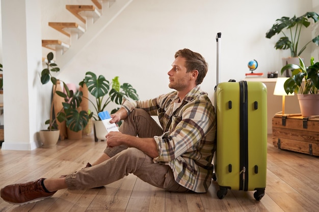 Portrait d'un beau touriste souriant tenant un passeport et des billets d'avion assis avec une valise