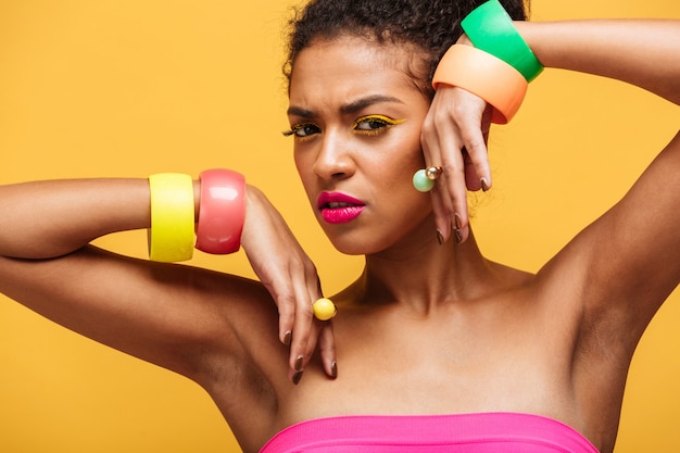 Portrait de beauté de la belle femme afro-américaine avec des cosmétiques colorés et des bijoux sur les mains posant avec un regard significatif isolé, sur le mur jaune