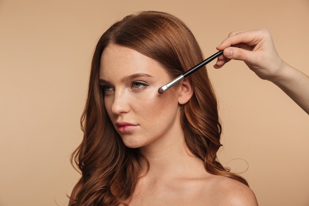 Portrait de beauté de femme au gingembre calme aux cheveux longs en détournant les yeux pendant que quelqu'un applique des cosmétiques avec une brosse
