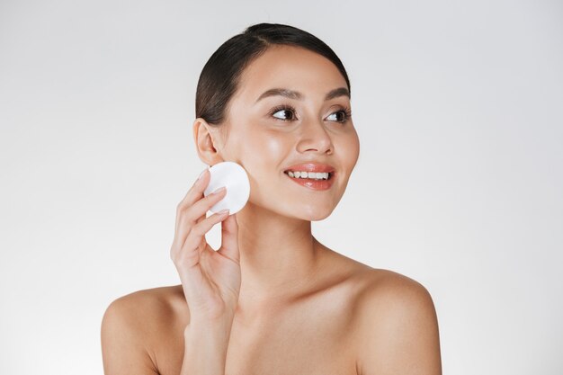 Portrait de beauté de femme brune souriante avec une peau saine et douce, démaquillant avec un coton, isolé sur blanc