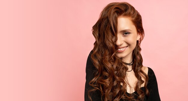 portrait de beauté d'une femme rousse posant sur un mur rose. Poils ondulés. Sourire parfait.