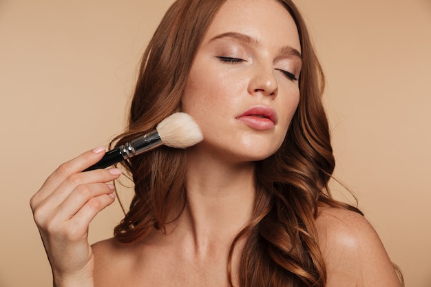 Portrait de beauté d'une femme sensuelle au gingembre aux cheveux longs et les yeux fermés en appliquant des cosmétiques avec une brosse