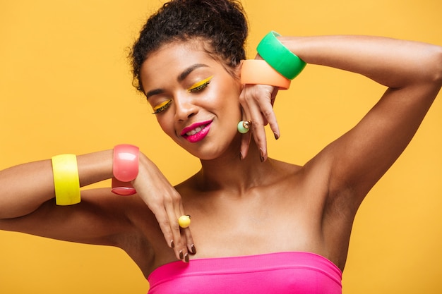Portrait de beauté de jolie femme afro-américaine avec maquillage mode et bijoux sur les mains posant isolé, sur mur jaune