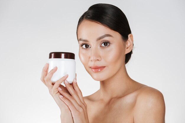 Portrait de beauté de la jolie jeune femme avec une peau douce et saine tenant la crème pour le visage hydratante et regardant la caméra, isolé sur blanc