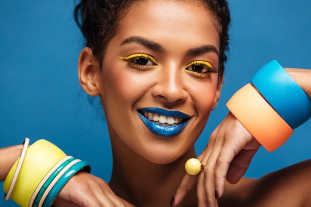 Portrait de beauté de la magnifique femme afro-américaine avec le maquillage de mode et les bracelets sur les mains regardant la caméra avec le sourire isolé, sur le mur bleu