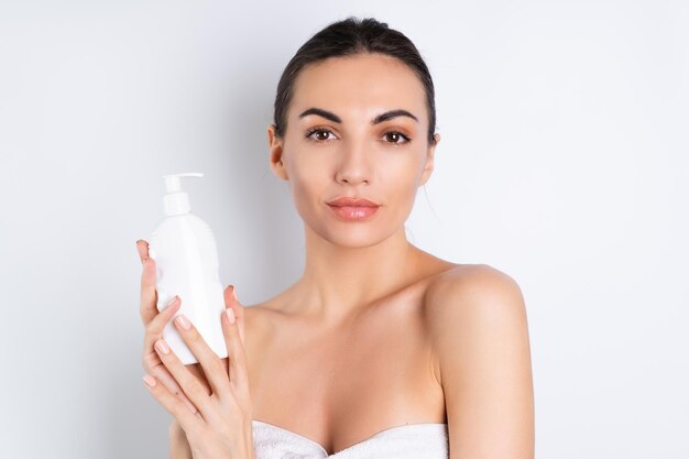 Portrait de beauté proche d'une femme aux seins nus avec une peau parfaite et un maquillage naturel tenant une bouteille de shampooing lotion pour le corps sur fond blancx9