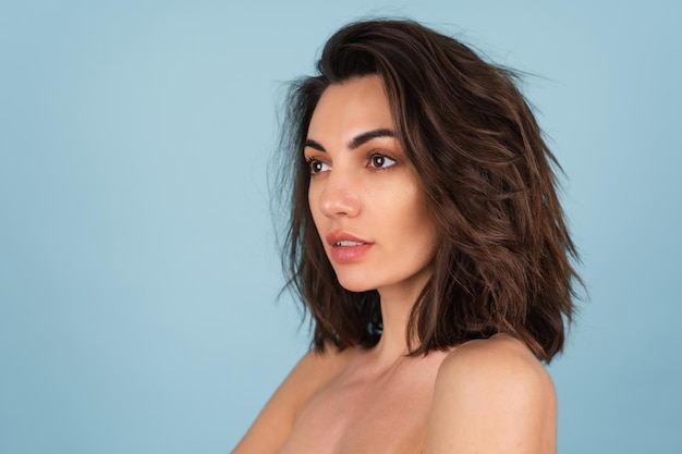 Portrait de beauté seins nus d'une jeune femme aux épaules nues et au maquillage naturel léger, aux cheveux courts ondulés, retouchée, sur fond bleu clair