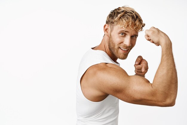 Portrait d'un bel athlète de fitness fléchissant les biceps montrant un bras musclé fort et un entraînement souriant dans une salle de sport debout sur fond blanc