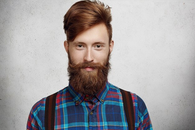 Portrait de bel étudiant avec barbe et moustache à la mode vêtu d'une chemise à carreaux à la mode avec une expression heureuse et joyeuse tout en se relaxant à l'intérieur après des conférences à l'université