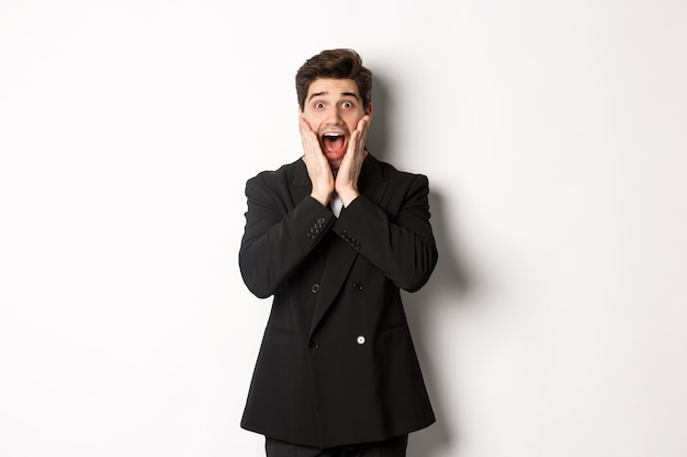 Portrait d'un bel homme d'affaires surpris en costume, réagissant à la promotion des vacances, semblant étonné de la caméra, debout sur fond blanc