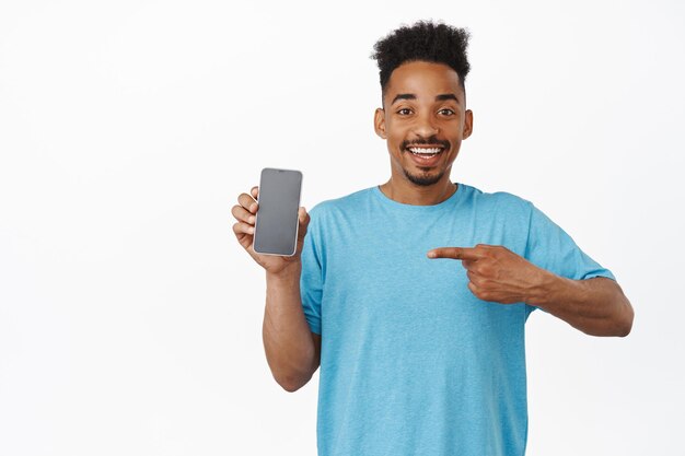 Portrait d'un bel homme afro-américain heureux pointant le doigt sur un téléphone portable, souriant heureux, montrant une publicité pour smartphone, recommandant un magasin ou une application, fond blanc