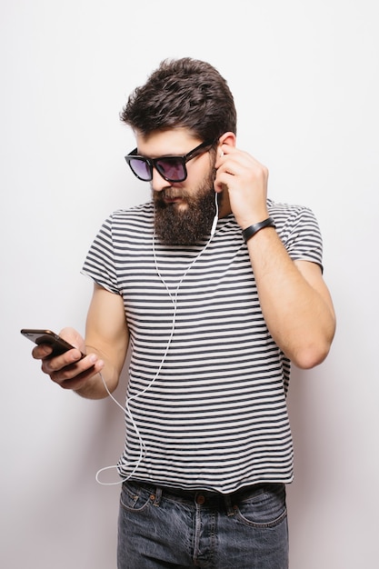 Portrait d'un bel homme barbu attrayant avec des écouteurs tenant un téléphone mobile isolé sur le mur blanc