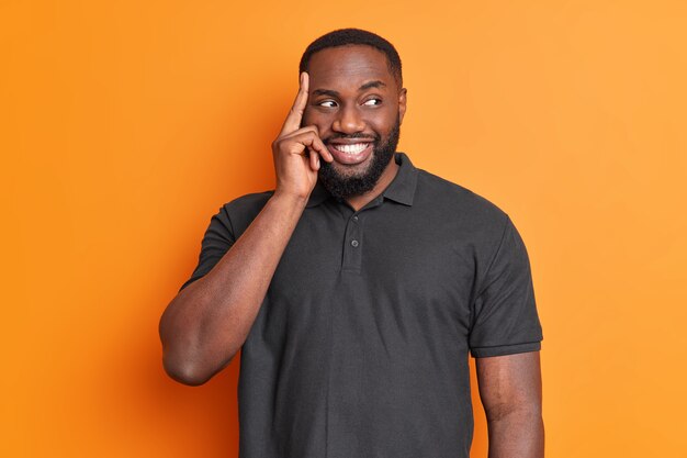 Portrait de bel homme barbu réfléchi garde le doigt sur le temple sourit pense agréablement à la décision regarde ailleurs habillé en t-shirt noir décontracté isolé sur mur orange