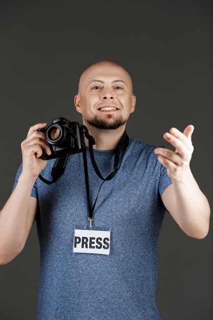 Portrait de bel homme en chemise grise avec photocamera et badge presse prendre des photos sur un mur sombre