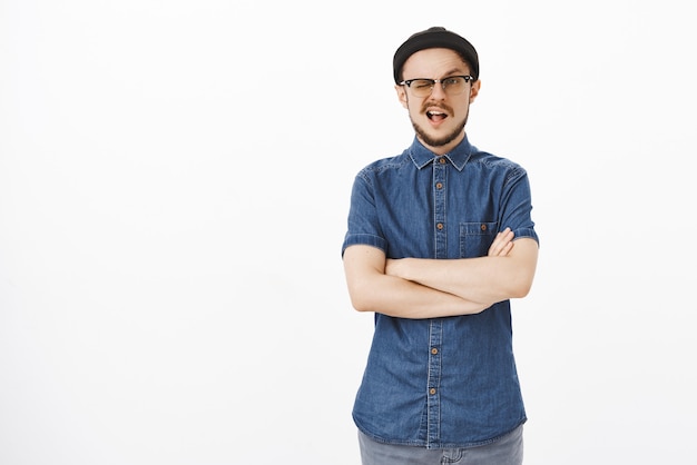 Portrait de bel homme confiant et élégant confiant en bonnet noir et lunettes avec barbe clignotant tenant les mains croisées sur la poitrine prête et accomplie